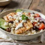 A vibrant Greek Chicken Bowl with grilled marinated chicken, rice, fresh cucumbers, cherry tomatoes, red onion, feta cheese, olives, and a drizzle of creamy tzatziki sauce.