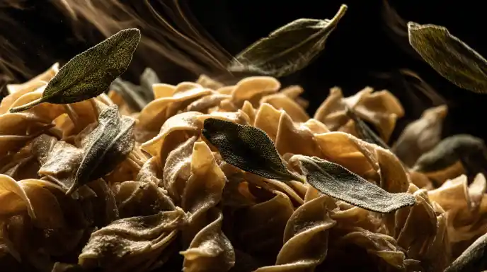 An action shot of freshly cooked garganelli pasta being tossed in a hot skillet with shimmering brown butter and crispy sage leaves. A wooden spoon is visible, and you can see the sauce coating each piece of pasta.